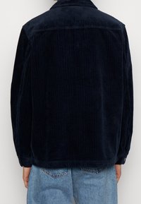 Chaqueta de pana azul marino con rayas verticales, que presenta un cuello, puños con botones y un corte relajado, combinada con jeans claros.