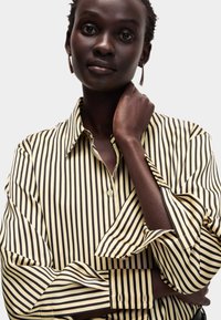 Camicia a righe in crema e nero, con colletto, maniche lunghe con polsini, e una vestibilità rilassata. Tessuto morbido, con una leggera lucentezza.