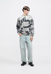 Jovem vestido com um moletom em tie-dye preto e cinza, jeans azuis claros, sapatos pretos e óculos de sol escuros, de pé com uma mão no bolso.