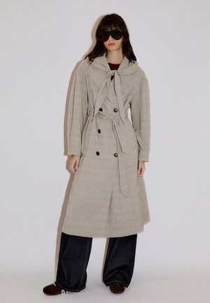 Femme portant un trench-coat beige oversize, pantalon large foncé, grandes lunettes de soleil noires, debout devant un fond uni.