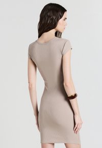 Robe bodycon beige à côtes avec des manches courtes, présentant un décolleté rond et une silhouette ajustée. Accompagnée d'un bracelet marron au poignet.