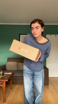 Barna kartondoboz narancssárga Zalando logóval, egy kék és fehér csíkos pólót viselő, világoskék farmernadrágos ember kezében, egy zöld fal előtt.