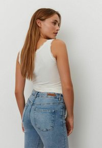 Vit linne med spetskant vid axeln och ljusblå hög midja jeans. Modellen poserar bakifrån, håret faller över ena axeln.