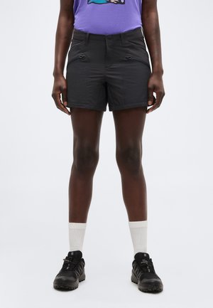 Άτομο που φοράει μαύρα σορτς με φερμουάρ, λευκές κάλτσες, μαύρα ορειβατικά παπούτσια και μωβ πουκάμισο με γραφικό σχέδιο.