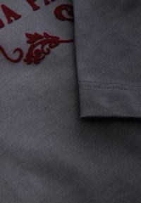 Grau Baumwollstoff mit glatter Textur, verziert mit burgunderroten gestickten Blumenmotiven und einem umgeschlagenen Ärmel, der saubere Nähte zeigt.