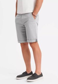 Shorts gris clair en coton, avec un ourlet roulé, des poches latérales et une texture lisse. Associés à des chaussures décontractées gris foncé.