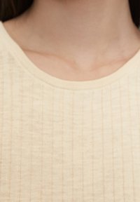 Cremefarvet, ribbestrikket top med rund hals. Glat overflade med subtile lodrette linjer, der fremhæver det lette stof.