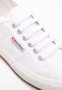 Bílý plátěný teniska se šesti průchody na tkaničky, stříbrným kováním, texturovaným povrchem a červeno-modrou štítkem s logem "Superga" na boku.