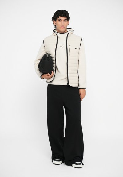 Beige quiltet vest med sorte detaljer, lange sorte bukser og sort veske, kombinert med en lyse farget topp og sorte sneakers.