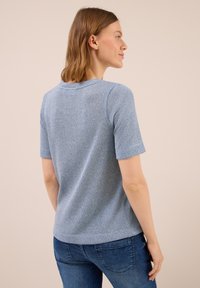 Frau mit hellbraunen Haaren, die nach rechts schaut, kleidet sich in einem kurzärmeligen, strukturierten hellblauen Oberteil und dunklen blauen Jeans vor einem beigen Hintergrund.
