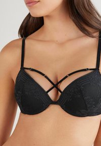 Soutien-gorge noir avec motif en dentelle, bonnets rembourrés, bretelles réglables et design en croix sur le devant. Tissu texturé avec un éclat subtil.