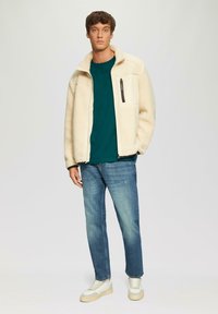 Fleece jack in crème, met ritssluiting aan de voorkant, hoge kraag, gestructureerd oppervlak, gecombineerd met een groene t-shirt, lichtblauwe jeans en witte sneakers.