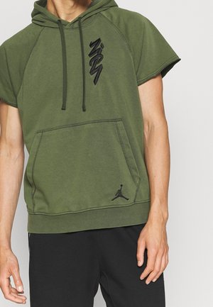 Man draagt een olijfgroene korte mouw hoodie met zwarte trekkoorden, een buidelzak en een zwart grafisch logo, gecombineerd met zwarte broek.