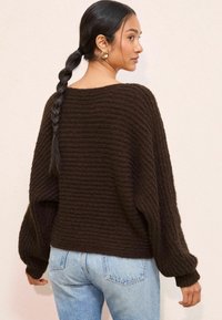 Brązowy sweter dzianinowy o luźnym kroju, szerokich rękawach i strukturalnym prążkowanym wzorze. Noszony z jasnoniebieskimi dżinsami.
