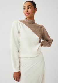 Maglione lavorato a maglia con colletto alto, caratterizzato da un design a blocchi di colore diagonali in crema e marrone. Texture morbida, maniche lunghe con dettaglio a palloncino.