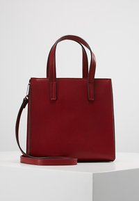 Punainen nahkakäsilaukku, jossa on rakenteellinen suorakulmainen muoto, kaksi lyhyttä kahvaa ja irrotettava olkahihna. Silkinen pinta ja minimaalinen muotoilu.