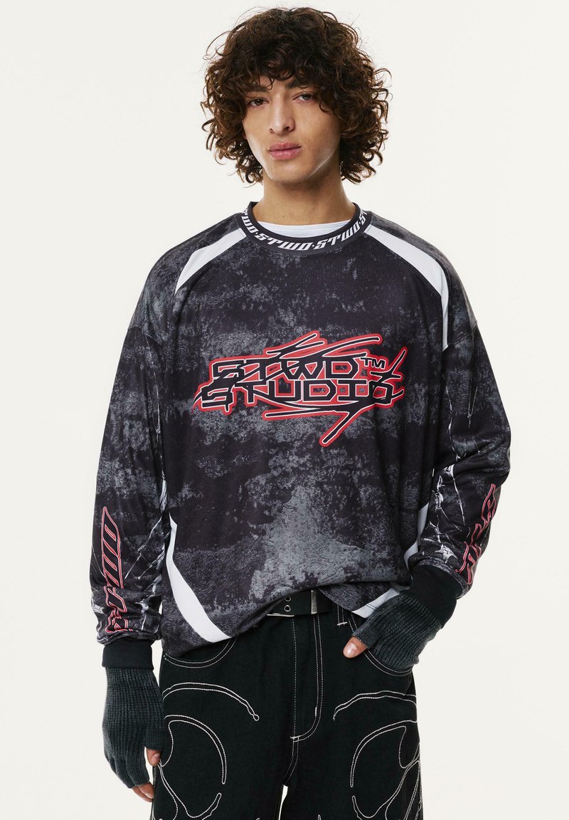 Črn pulover z teksturiranim, abstraktnim vzorcem, ki vsebuje bele poudarke in rdeč grafični logo. Ima dolge rokave in teksturo brezprstnih rokavic.