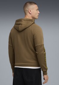 Mand, der står sidelæns iført en brun hættetrøje med mønstrede bånd på ærmet og sorte bukser mod en grå baggrund.