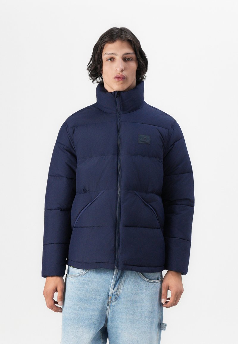 Marineblauwe pufferjack met hoge kraag, ritssluiting en twee voorzakken. De stof heeft een gladde textuur en een quiltpatroon.