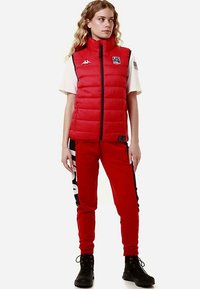 Gilet imbottito rosso con collo alto e zip nera, abbinato a pantaloni della tuta rossi con logo bianco e dettagli laterali neri. Stivali neri.