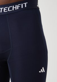 Leggings de sport bleu marine avec une taille élastique blanche "TECHFIT" et un logo à trois rayures blanches. Texture de tissu lisse, design ajusté.