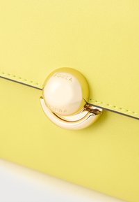 Żółta torebka ze skóry o fakturze, z okrągłym złotym zapięciem, z wytłoczonym logo na zapięciu oraz kontrastowym przeszyciem krawędzi.