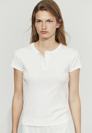 Frau mit langen braunen Haaren, die ein weißes Henley-Shirt mit kurzen Ärmeln trägt und vor einem einfarbigen hellen Hintergrund steht.
