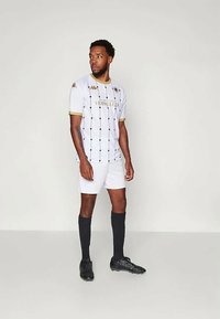 Λευκή αθλητική φανέλα ποδοσφαίρου με χρυσές λεπτομέρειες. Μπλούζα κοντού μανικιού με κάθετες μαύρες ρίγες και έμβλημα. Μαύρες κάλτσες και ποδοσφαιρικά παπούτσια ολοκληρώνουν την εμφάνιση.