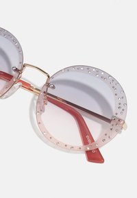Occhiali da sole rotondi con una montatura rosa con sfumature decorata con strass trasparenti, dettagli dorati e bracci trasparenti di colore rossastro.
