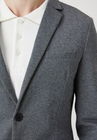 Blazer grigio con una trama strutturata, un unico bottone e una tasca sul petto, abbinato a una camicia bianca con colletto e bottoni sotto.