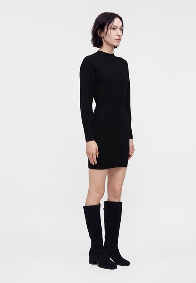 Femme portant une robe noire moulante à manches longues et des bottes noires hautes jusqu'aux genoux, debout devant un fond clair uni.