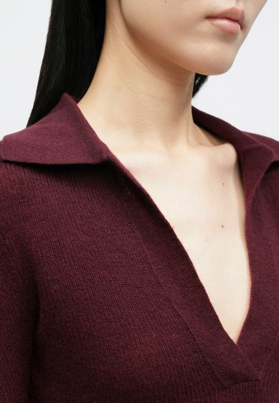 Κοντινό πλάνο ενός ατόμου που φοράει μπουκλωτό πουλόβερ σε βαθύ V λαιμόκοψη, μπορντό χρώμα, εμφανίζοντας τον λαιμό, τις κλείδες και το κάτω μέρος του προσώπου με ουδέτερο χρώμα χειλιών.