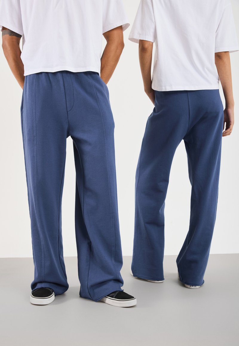 Pantaloni blu a gamba larga realizzati in tessuto morbido, con tasche laterali e una vestibilità rilassata. Abbinati a sneakers nere casual e camicie bianche corte.