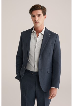 Blazer marine avec un motif texturé, fermeture à un bouton, revers notched, et une chemise boutonnée de couleur claire en dessous. Coupe ajustée.