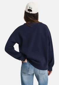 Marineblå overdimensjonert sweatshirt med lange ermer, ribbede mansjetter og rund hals, kombinert med lyseblå jeans. Hvit caps med grønn tekst.