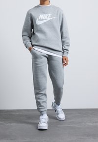 Sudadera gris y pantalones jogger de tejido suave, con un logo de Nike en blanco. Se complementa con zapatillas blancas y una camiseta blanca de varias capas debajo.