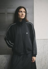 Giacca nera con zip realizzata in tessuto liscio, con strisce laterali bianche. Collo alto, vestibilità comoda e dettagli con polsini e orlo a coste.