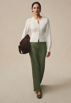 Femme portant un cardigan blanc boutonné, jupe vert olive, tenant un sac marron foncé, marchant vers l'avant sur un fond beige uni.