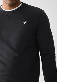 Man wearing a black long-sleeve sweatshirt with a small white bird emblem on the chest, layered over a white crew-neck shirt.