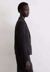 Black pinstripe blazer with a notched collar, two buttons, and pockets. Textured fabric with a structured fit. Model shown in profile.