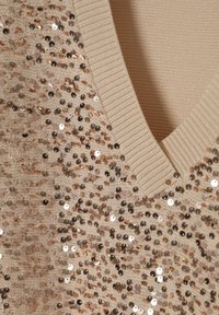 Bēša adīta virskrekls ar V veida izgriezumu, rotāts ar izkaisītiem rozā zelta spīdumiem, un ribota kakla daļa. Gluda tekstūra ar mirdzumu.