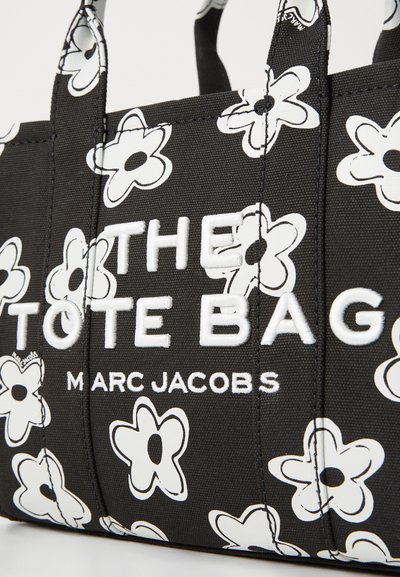 Sac fourre-tout noir avec un motif floral blanc, présentant un texte blanc en évidence : "THE TOTE BAG M A RC JACOBS," fabriqué en tissu durable avec des poignées solides.