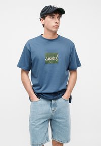 Νέος άνδρας που φοράει μπλε μπλουζάκι Rip Curl, ανοιχτόχρωμα τζιν σορτς και καπέλο με ασπρόμαυρες ρίγες, στέκεται με τα χέρια στις τσέπες.