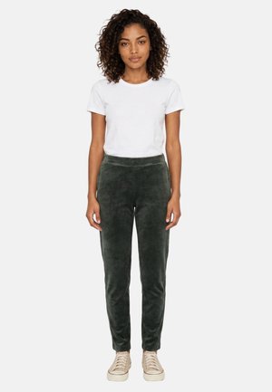 Joven mujer con pelo rizado que lleva una camiseta blanca, pantalones de terciopelo verde oscuro y zapatillas beige, de pie contra un fondo claro.