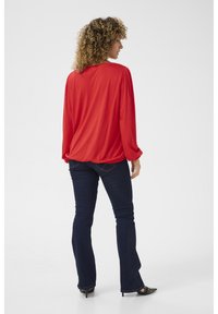Rotes lang�ärmliges Oberteil mit gerafften Schulterdetails, kombiniert mit dunkelblauen ausgestellten Jeans. Glatter Stoff, lockere Passform und seitliche Saumakzente.
