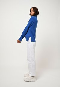 Blauer Cashmere-Turtleneck-Pullover mit seitlichen Schlitzen, kombiniert mit weißen Jeans und beigen chunky Sneakers. Einfaches, sauberes Design auf neutralem Hintergrund.