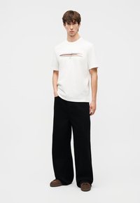 Fiatal férfi áll, fehér pólót visel, amelyen többszínű vízszintes csík található, bő fekete nadrágot, barna cipőt hord, kezét a zsebében tartja.