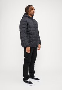 Black puffer jacket with a hood, featuring horizontal quilting, zip pockets, and elastic cuffs, paired with black pants and black sneakers.