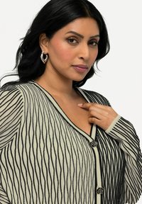 Musta ja kerma raidallinen neuletakki V-kaula-aukolla, hopeisilla ruusupainikkeilla ja teksturoidulla kankaalla, jossa on pitkät hihat ja vartalonmyötäinen muotoilu.