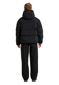 Svart puffjacka med huva, med kviltade sektioner. Bärs över svarta byxor, som visar en strömlinjeformad, isolerad design.